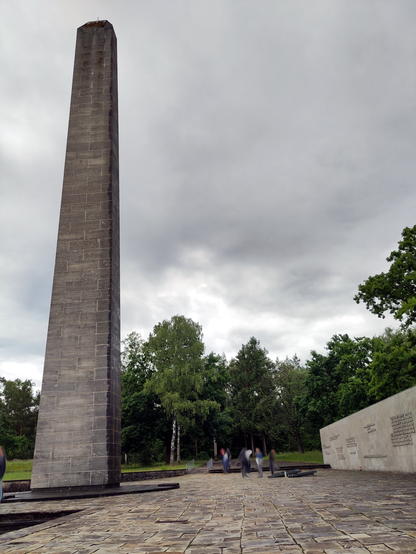 Der Obelisk auf der Gedenkstätte der Gedenkstätte Bergen-Belsen, drumherum ein mit Naturstein gefpalsterter Platz und dahinter eine Gedenkwand mit Gedenksprüchen in verschiedensten Sprachen.