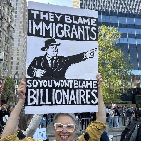 A smiling protester holds a large illustrated sign at a public demonstration in a city square surrounded by tall buildings. The sign features a black-and-white drawing of a man in a suit and hat pointing outward, with bold text that reads: “THEY BLAME IMMIGRANTS SO YOU WON’T BLAME BILLIONAIRES.” The style of the sign resembles vintage political posters. A crowd of fellow demonstrators is visible behind metal barricades in the background.