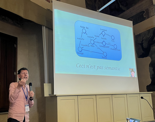 Frank standing on the left side next to the projection screen showing a labelled, directed graph. The graph is captioned by the sentence "Ceci n'est pas sèmantic."