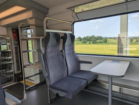 Zwei leere Zugplätze an einem hellen Holztisch blicken durch ein großes Fenster auf grüne Felder und Bäume unter blauem Himmel. Das Interieur ist modern mit Gepäckablagen und grauen Polstern.