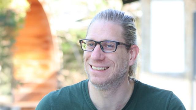 Porträt von Rainer Prüßmann. Ein lächelnder Mann mit langen, blonden, nach hinten gebundenen Haaren und Brille. Er trägt ein dunkelgrünes T-Shirt und steht vor einem unscharfen Hintergrund.