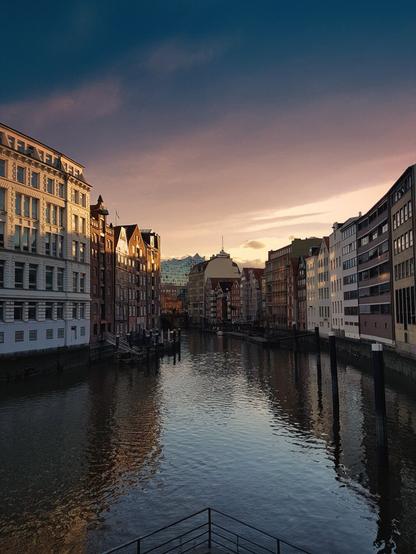 A wide shot of a canal in Hamburg at sunset, flanked by tall, historic European buildings on both sides. The sky above transitions from a deep blue at the top to soft purples and oranges near the horizon, reflecting gently on the calm water of the canal. The buildings on the left are predominantly light-coloured with many windows, while those on the right are darker, some with traditional gabled roofs. In the distance, a modern, distinctively shaped building with a wave-like roof can be seen. The overall mood is serene and picturesque.
