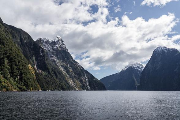 View of Milford Sound (Piopiotahi) with steep fjord walls and snow-capped peaks under a partly cloudy sky, captured from a boat on the water.