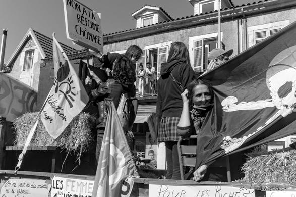 Demonstrators mounted straw bales on a vehicle bearing slogans against sexism and unemployment insurance reform, as well as a pirate flag and a farmers' union flag. Behind, a couple watch the demonstrators from their balcony.