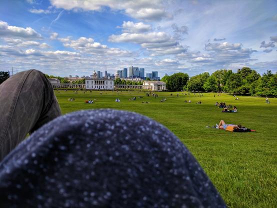 A first-person perspective shows two people relaxing in Greenwich Park, London. One person's legs, clad in dark trousers, and the other person's pregnant belly, dressed in a dark grey t-shirt, are visible in the foreground, slightly out of focus. Beyond them, a wide, sunlit green lawn stretches into the distance, dotted with numerous people picnicking and lounging. In the middle ground, white classical buildings with columns are visible, and further back, the modern skyscrapers of Canary Wharf rise under a bright blue sky with fluffy white clouds. Lush green trees line the right side of the park.