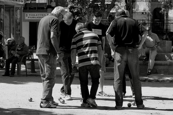 ein schwarz-weiß foto zeigt eine gruppe älterer männer, die sich über kugeln, wahrscheinlich pétanque-kugeln, im sand beugen und diskutieren.
sie sind auf einem platz in einer korsischen ortschaft. im hintergrund sitzen ältere männer. die szene wirkt entspannt und gesellig. der ton ist dokumentarisch und alltäglich. die szene strahlt eine ruhige, fast meditative atmosphäre aus. die kontraste zwischen licht und schatten betonen die texturen der kleidung und des bodens.