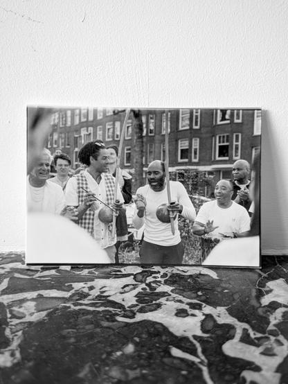 A printed black and white photo of a capoeira performance. Two men are playing the traditional berimbau instrument, other men are clapping rhythms with their hands.