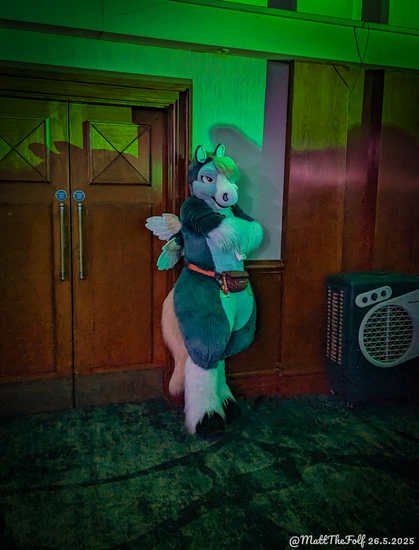 This is a candid photograph of a blue pegasus fursuiter standing off to the side of the main dance floor at ConFuzzled 2025. He's got his arms crossed in an imposing manner with green lighting and the silhouettes of other dancers washing over him. He might look a little intimidating but he's really just trying to cool down next to a fan that isn't blowing nearly as hard as it ought to be.