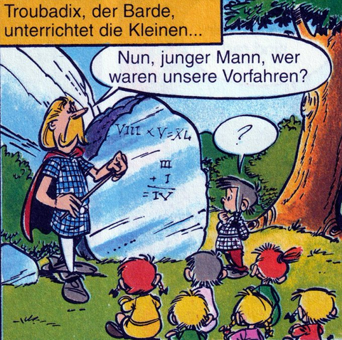Troubadix steht als Lehrer vor jungen Schülern. Hinter ihm ein Steinblock, der als Tafel genutzt wird. Ein Schüler steht neben ihm.