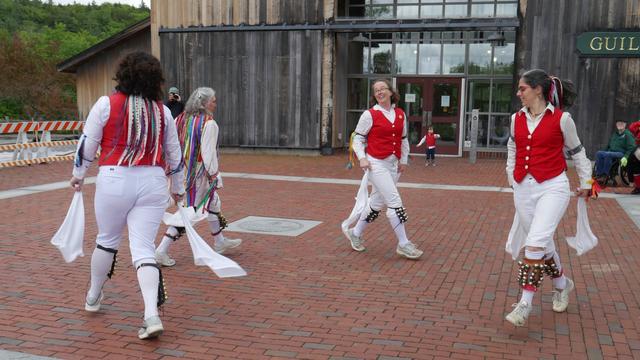 Four morris dancers with white shirts, knee britches, and stockings and red vests, decorated with many multi color ribbons, dancing on a brick paved surface. They are moving in a circle, hands low. There are some audience members behind them.