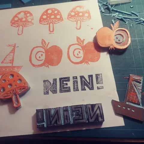 Ein Stück Papier mit Stempel-Probedrucken und den jeweiligen Stempeln: Ein Fliegenpilz, ein Apfel und ein Segelboot in orange, das Wort "Nein" in Großbuchstaben in schwarz.