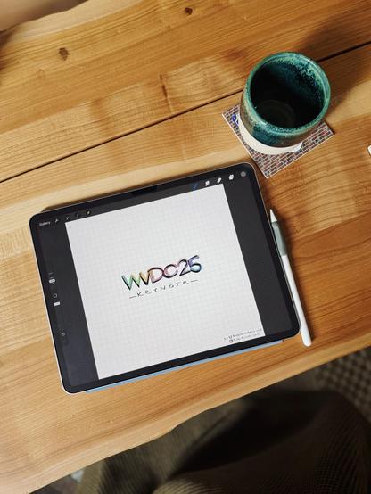 An iPad on a wooden surface, displaying a colorful "WWDC25 Keynote" design on a grid background. Next to the iPad, there is an Apple Pencil and a ceramic mug