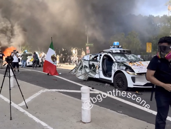 Screenshot uit video @bgonthescene: Vernielde en brandende Waymo robot-taxi’s bij rellen in straten van Los Angeles. Oa een Mexicaanse vlag is te zien en gemaskerde demonstrant/fotograaf.
