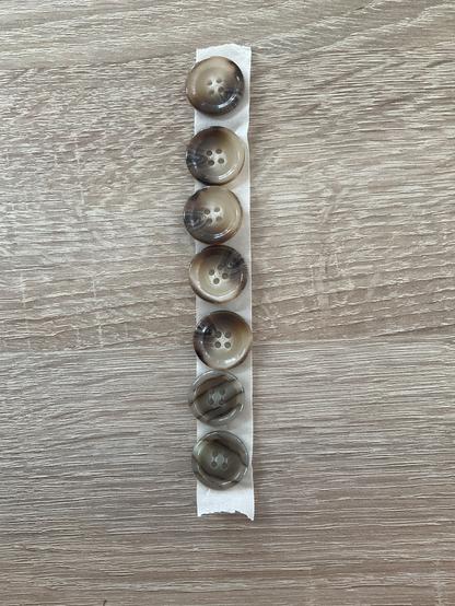 Das Bild zeigt eine Reihe von sechs Knöpfen, die auf einem weißen Malerkrep angeordnet sind. Dieses liegt auf einer eichenfarbenen Tischplatte