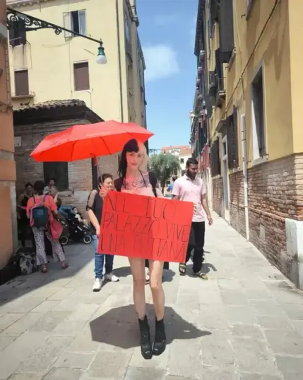 L'immagine mostra una giovane donna con capelli bicolore (neri e bianchi) che tiene un cartello rosso in una stretta strada di Venezia. Il cartello recita "Nel tuo palazzo vive una puttana". La donna indossa un vestito corto e stivali neri. Tiene un ombrello rosso. Sullo sfondo ci sono altre persone, alcune delle quali sembrano turisti.