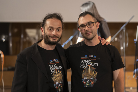 Umberto Ugoberti (a sinistra) e Nicola Monopoli (a destra), docenti della Scuola di Musica Elettronica del Conservatorio di Foggia, posano insieme durante l’evento “Sonic Explorations”, indossando le magliette ufficiali del Blended Intensive Programme Erasmus. Sullo sfondo, palco e microfoni sfocati.