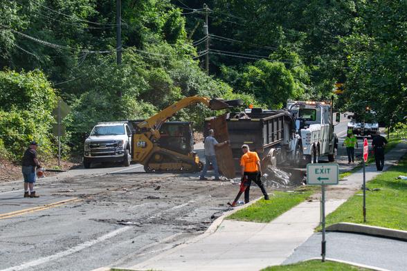 Small yellow front-loader dumping a tire into a large waste truck that is ready to be towed away. Men with leaf blowers are in the foreground trying to look busy.