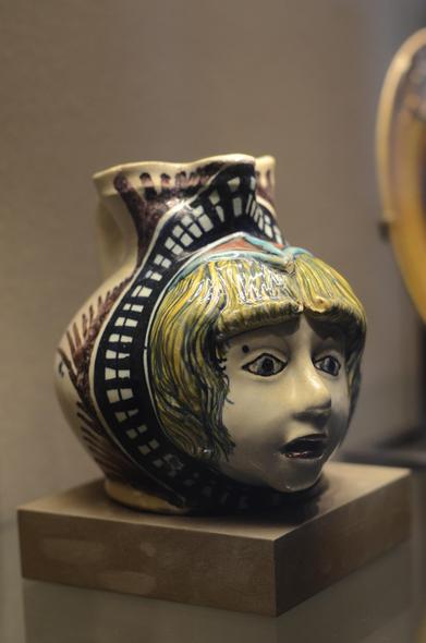 A wide pitcher glazed in many colors, with the face of a boy sculpted on the belly. He's got yellow hair and a bit of a panicky look on his face.