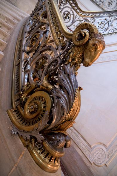 Décoration très chargée du bas de la rampe d’un escalier. Une tête de bélier en métal, des feuilles forgées…
