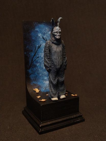 Das Bild zeigt eine detaillierte Figur in einem bunten, tierähnlichen Kostüm, die auf einem schwarzen Podest steht. Die Figur hat lange Ohren und ein maskenhaftes Gesicht mit einem gruseligen Lächeln. Sie trägt eine dunkelgraue, rissige Textur, die an Fell erinnert. Die Figur steht auf einem schwarzen Podest, das mit einigen gelben Blättern verziert ist. Im Hintergrund ist eine künstlerische Darstellung eines blauen Nachthimmels mit dunklen Ästen zu sehen. Die Figur ist in einer ruhigen, aber mysteriösen Pose dargestellt, was durch die dunkle Umgebung und die künstlerische Hintergrundgestaltung verstärkt wird.