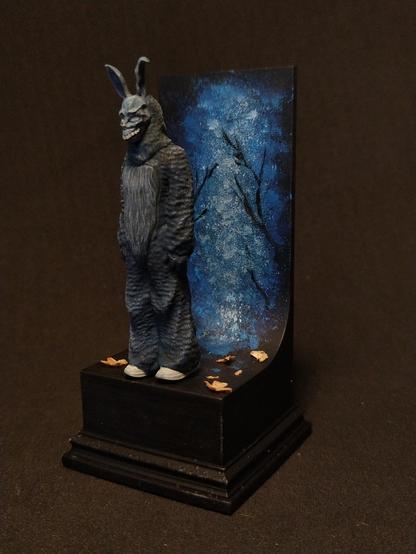 Das Bild zeigt eine detaillierte Figur in einem bunten, tierähnlichen Kostüm, die auf einem schwarzen Podest steht. Die Figur hat lange Ohren und ein maskenhaftes Gesicht mit einem gruseligen Lächeln. Sie trägt eine dunkelgraue, rissige Textur, die an Fell erinnert. Die Figur steht auf einem schwarzen Podest, das mit einigen gelben Blättern verziert ist. Im Hintergrund ist eine künstlerische Darstellung eines blauen Nachthimmels mit dunklen Ästen zu sehen. Die Figur ist in einer ruhigen, aber mysteriösen Pose dargestellt, was durch die dunkle Umgebung und die künstlerische Hintergrundgestaltung verstärkt wird.