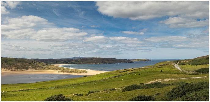 A panoramic view of a coastal landscape featuring sandy beaches, a winding river, rolling green hills, and scattered clouds in a blue sky. A road runs through the scene, leading towards a distant white house.