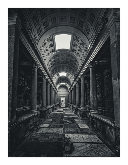 This photo is of a black and white photograph of a long, dimly lit hallway/padsageay. The hallway is lined with columns and alcoves, which contain plaques for tombstones/burial spaces. There are located in the flooring and in the walls. The architecture is classical, with an arched ceiling with lots of intricate details. The perspective leads the eye down the length of the corridor to a distant light source - a brightly lit doorway that leads into another room.
