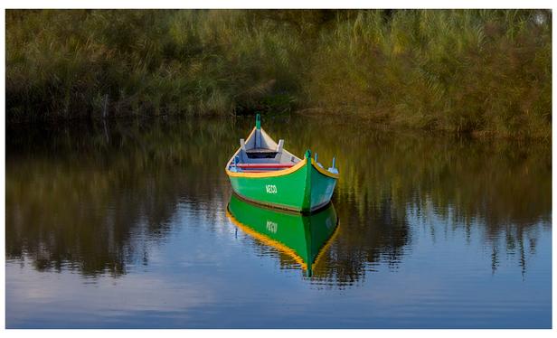 Barquiña de cores vivos nunha lagoa de augas mansas que reflicten un ceo azul con algunha nube. (galego)

A brightly coloured boat on a lake with calm waters reflecting a blue sky with some clouds.