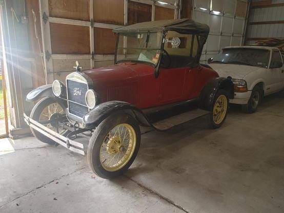 Front driver side corner view of a 1927 Model T Runabout. Red body, black fenders, and straw colored wire wheels.