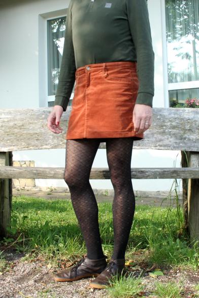 Das Bild zeigt eine stehende Person im Freien, deren Kopf nicht sichtbar ist. Die Person trägt ein langärmliges, olivgrünes Oberteil, einen kurzen, orangefarbenen Cordrock, schwarze gemusterte Strumpfhosen und braune Lederschuhe mit Schnürung. Im Hintergrund sieht man eine Hauswand mit Fenstern sowie eine einfache Holzbank auf einem mit Gras und Kies bedeckten Boden.