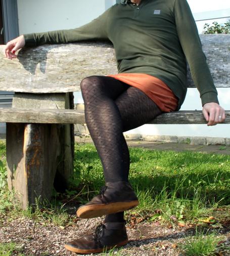 Das Bild zeigt eine sitzende Person auf einer rustikalen Holzbank im Freien. Die Person trägt ein olivgrünes, langärmliges Oberteil, einen kurzen, rostorangefarbenen Cordrock sowie schwarze, gemusterte Strumpfhosen und dunkelbraune Schnürschuhe mit Gummisohle. Ein Bein ist locker über das andere geschlagen, die Haltung wirkt entspannt.