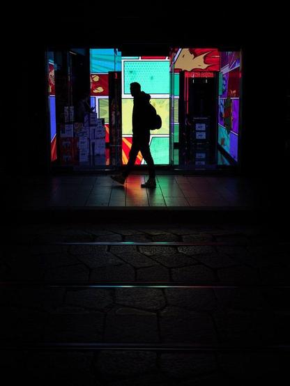 The silhouette of a man with a backpack walking past a shop window at night. The inside of the shop is illuminated by several colorful light panels, creating a wide range of colors.