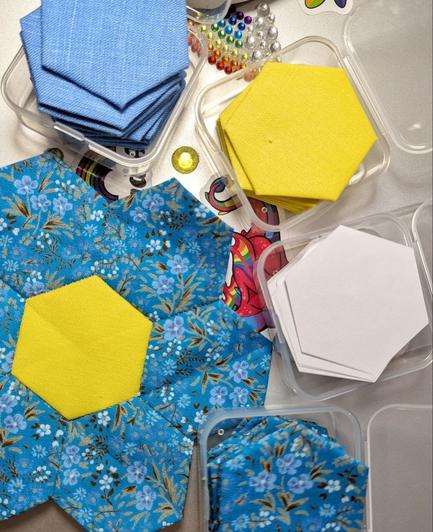 Fertig genähte Hexagon-Blüte für den Flowergarden Quilt aus blauem Stoff mit kleinen Blümchen und einem gelbem Mittelstück. Runherum durchsichtige kleine Plastikdöschen mit zum Verarbeiten bezogenen Hexagons in gelb, blau, blauen Blümchen und ein Kästchen mit Papierschablonen für weitere Projekte.