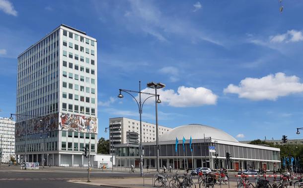 In der linken Bildhälfte ist das Haus des Lehrers zu sehen. Ein graues Hochhaus mit einem umlaufenden bunten Mosaik etwas in der 3. und 4. Etage. Daneben ein flaches Gebäude mit einer Glasfassade und einer Kuppel
