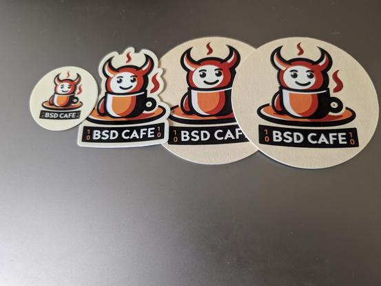 Two coasters and two stickers from "BSD Cafe" are arranged on a metallic gray surface.