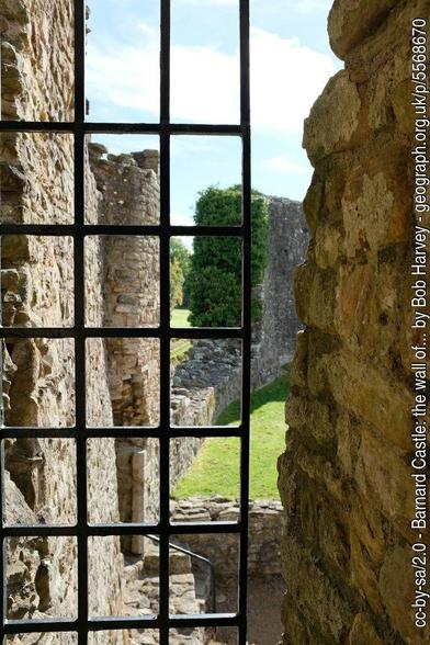 View through an iron grating of the gap between a castle curtain wall and the keep. Stone walls curving away from the foreground grating, a patch of grass and a blue sky in the distance