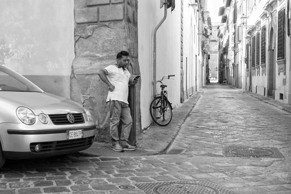Black and white photo of a man in a white t-shirt looking at his phone beside the front of a compact car. To the right, a narrow street stretches off into the distance. A bicycle lands against the wall.