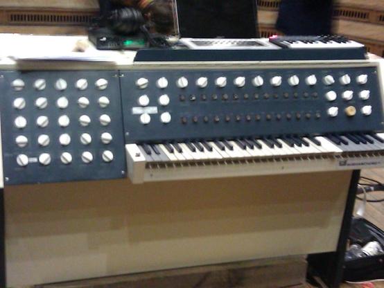 Mein unscharfes Foto des Subharchords aus der Nähe. Auf einem Korpus aus hellem Holz sind eine Klaviatur und ein großes Schaltpaneel aufgesetzt. 
Die Klaviatur hat am oberen und am unteren Ende Abschnitte, in denen die hellen Tasten in einem Grauton statt Weiß ausgeführt sind.
Das Schaltpannel besteht aus zwei blaugrauen Platten mit vielen weißen Drehreglern und Schaltern.
Eine Platte ist direkt vertikal über der Klaviatur angebracht.
Die zweite Platte ist links versetzt neben der Klaviatur angebracht.
Insgesamt wirkt die Gestaltung auf mich wie aus einem alten Science-Fiction-Film.