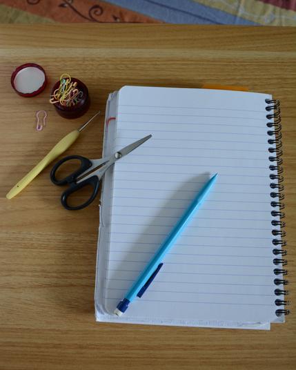 Carnet à spirales ouvert sur une page blanche, posé sur une table en bois clair, accompagné d'un crayon à papier bleu, d'une paire de ciseaux, d'un crochet et de quelques marqueurs de maille colorés.