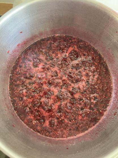 Vue en plongée sur une casserole en inoxydable grise où cuit de la confiture de framboises (rouge très foncé avec une sorte d’écume rose foncé)