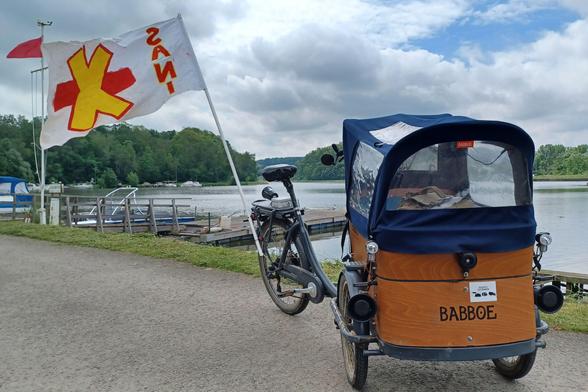 Ein Lastenfahrrad mit einer Kiste aus Holz steht bei leicht bewölktem Himmel vor einem Fluss, dessen anderes Ufer bewaldet ist. Daran eine lange Stange mit einer wehenden Fahne, auf der in roten Großbuchstaben “SANI” steht. An dem Lastenfahrrad sind kleine Lautsprecher montiert.