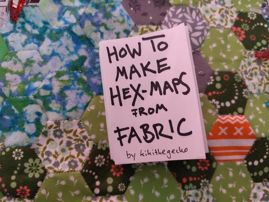 Handwritten zine "how to make hex maps from fabric by kikithegecko" that lays on a quilted hex map made from green and blue hexes