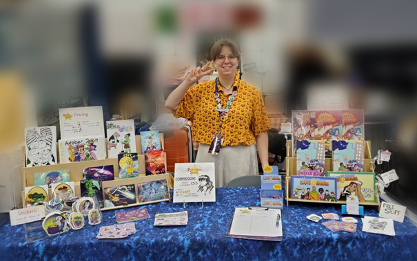 A smiling person behind a table full of comics, stickers, and prints.