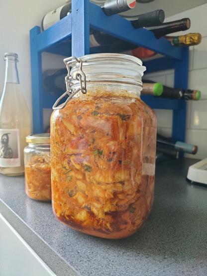 En stor och en liten burk med kimchi på en köksbänk framför ett blått vinställ med några flaskor i