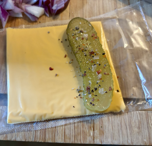 A slice of pickled gherkin adorned with a sprinkle of sea salt and peppers inside a slice of processed cheese ready to be folded over.