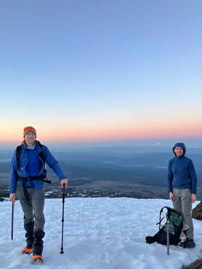 Two men with mountaineering gear smile at the camera while standing on snow. A peak is visible on the hazy orange horizon.