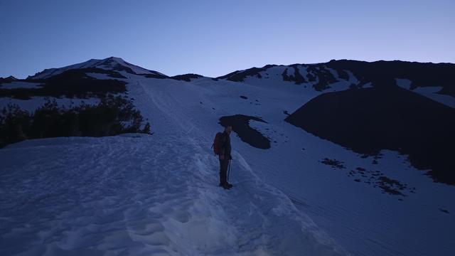 A mountaineer stands on a snowy ridge in predawn light.