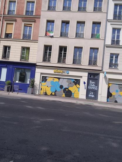 Photo d'une immeuble parisien. Au 2eme étage un drapeau palestinien et en dessous, au niveau de la rue, un immense graffiti en blanc sur une porte dit FAN CLUB HAMAS 2ÈME ÉTAGE avec une immense flèche bleue indiquant la fenêtre
