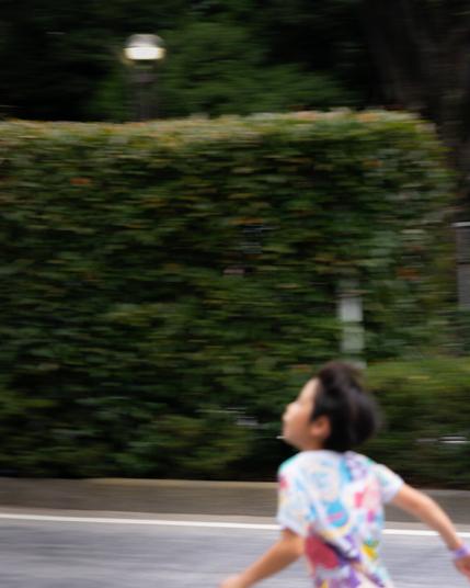 Blurred image of a child running on a pavement next to a lush, green hedge. The child's colorful shirt contrasts with the greenery. A dimly lit streetlamp is visible in the background, surrounded by dense foliage. The movement creates a sense of motion.