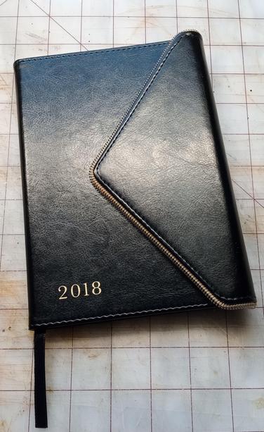 A black faux leather 2018 date book with a magnetic closure that wraps around the right side shaped like an envelope flap. The flap is trimmed in gold zipper teeth. There is a black ribbon coming from the binding. There is gold print on the lower left side that reads "2018."